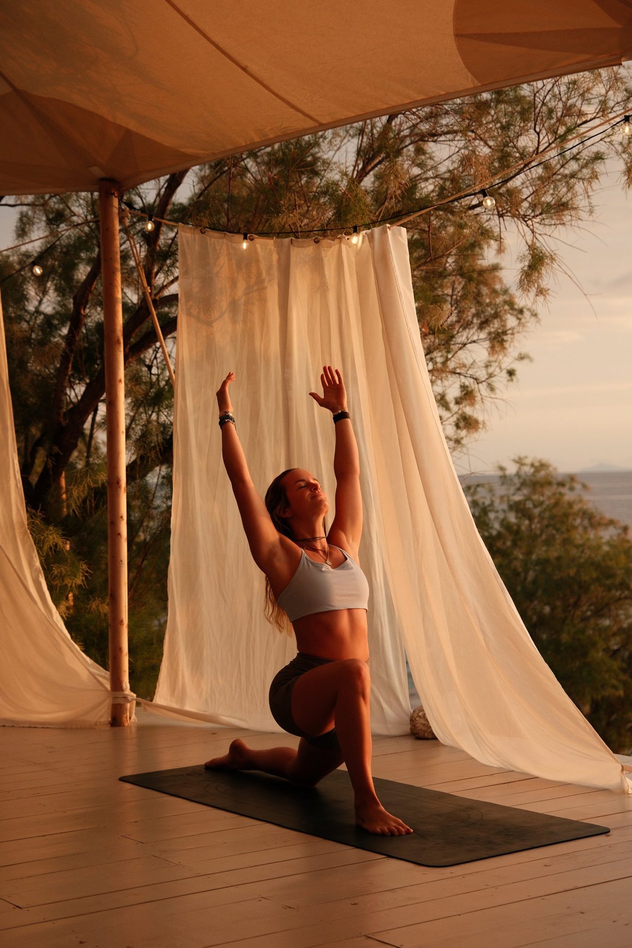 Tamara in a kneeling stretch with arms raised, at sunset under a canopy