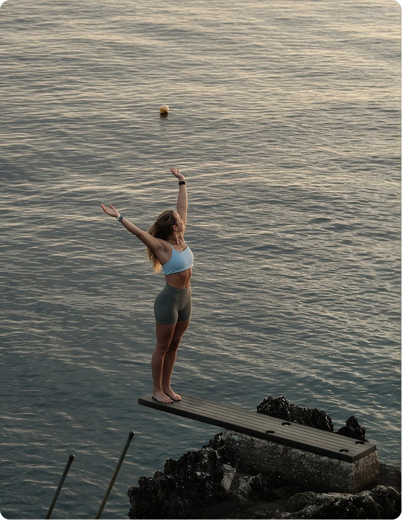 Tamara on a diving board above the sea at sunset, arms raised